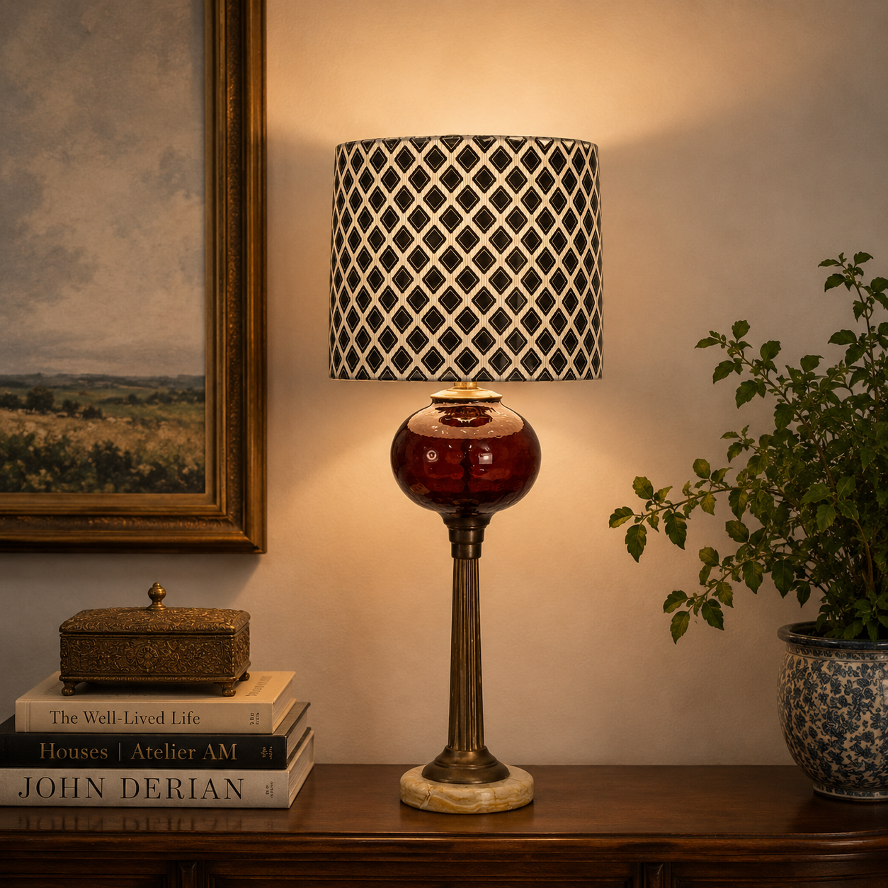 A table lamp with a black and white diamond-patterned drum shade, a glossy dark red glass bulb, and a fluted brass-toned stem, shown lit on a wooden console table in a styled room setting.