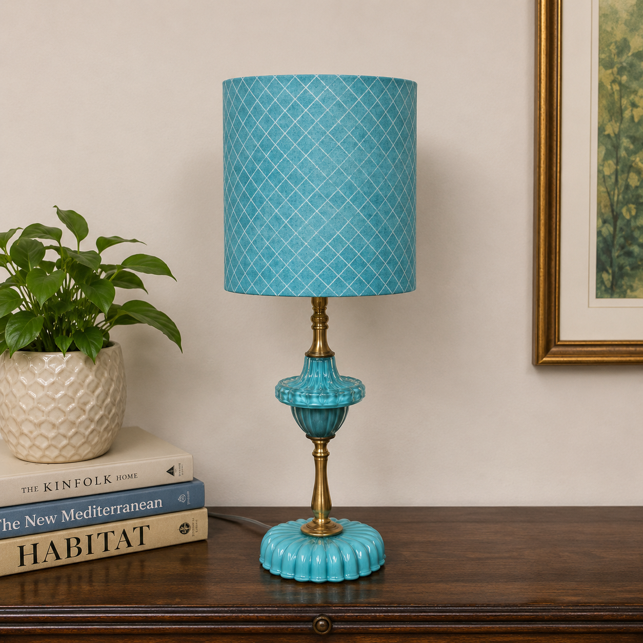 A turquoise and gold table lamp with a patterned blue-gray shade sits on a dark wooden console table next to a potted plant and books, with a framed picture on the wall in the background.