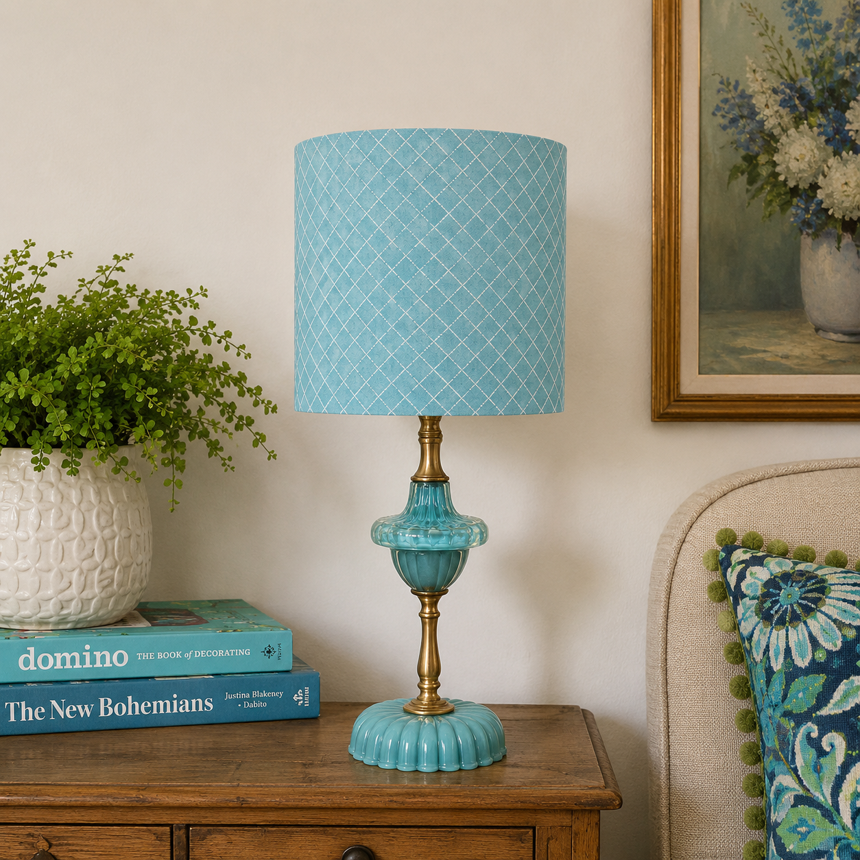 A turquoise blue table lamp with a patterned drum shade and fluted base, styled on a wooden side table in a cozy room setting next to a potted plant and a patterned pillow.