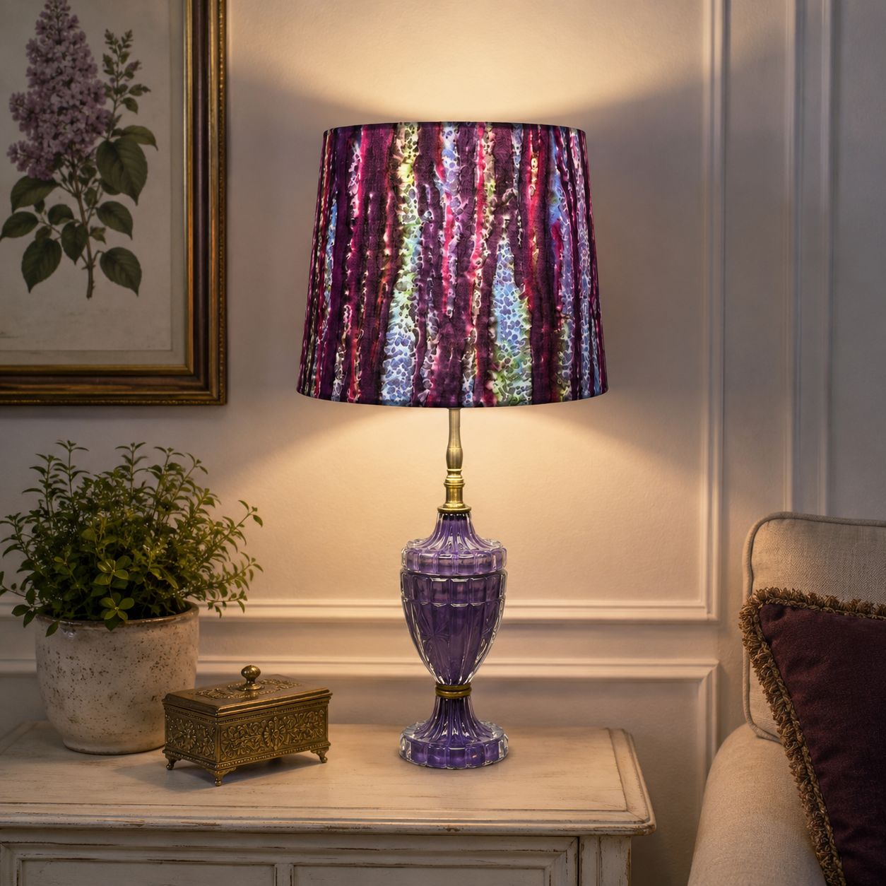 A table lamp with a purple glass base and a pleated fabric shade featuring a vertical iridescent stripe pattern in purple, pink, and blue, styled on a console table in a living room setting.